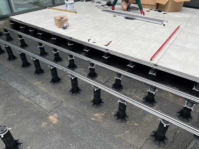 Instalación de una terraza elevada exterior utilizando plots regulables de plástico y vigas de aluminio portantes con baldosas cerámicas.