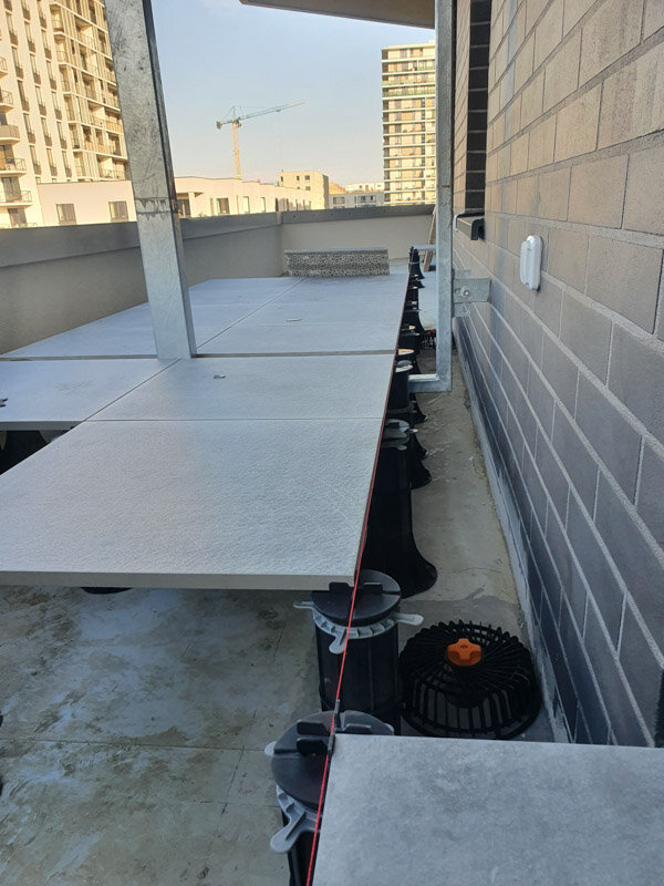 Línea láser roja de nivelación alineando baldosas de gres sobre soportes de terraza regulables altos a lo largo de la pared del edificio.