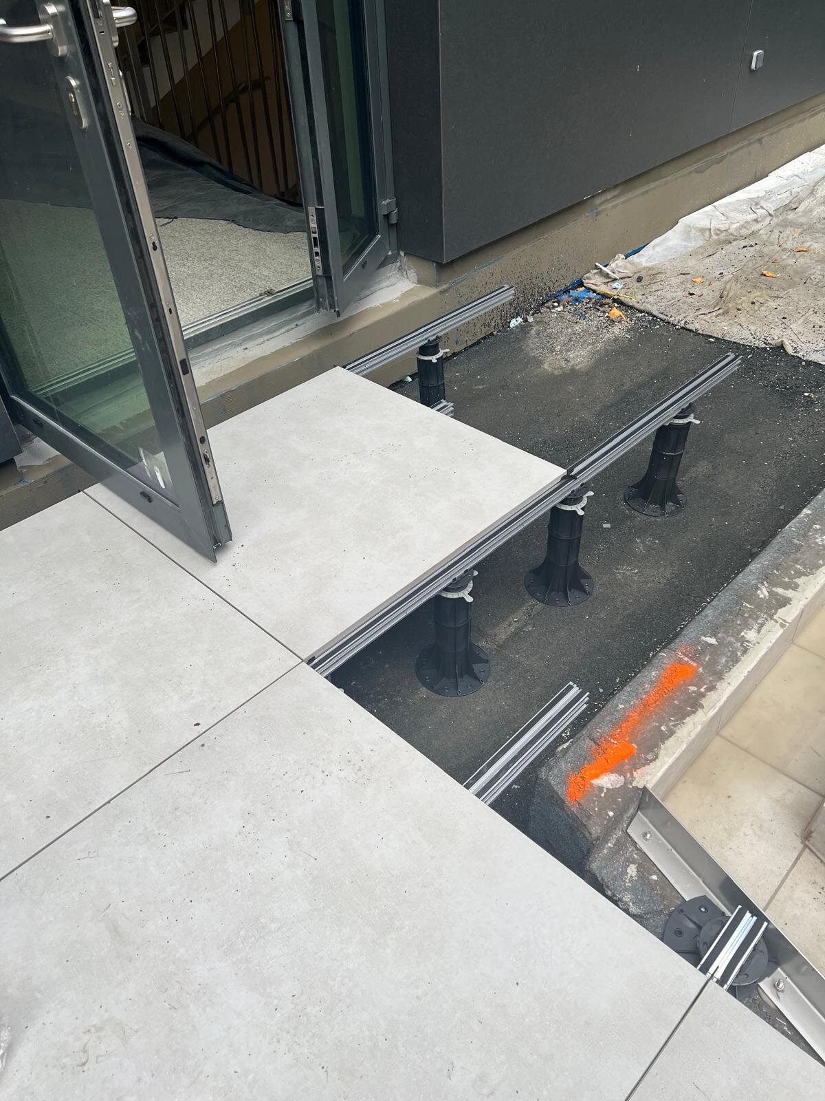 Instalación junto a una puerta de terraza con baldosas de porcelana para exterior colocadas sobre vigas de aluminio y plots soporte regulables de plástico para terrazas.