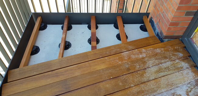 Instalación de tarima de balcón con vigas de madera visibles apoyadas sobre plots soporte regulables de terraza sobre base de hormigón.