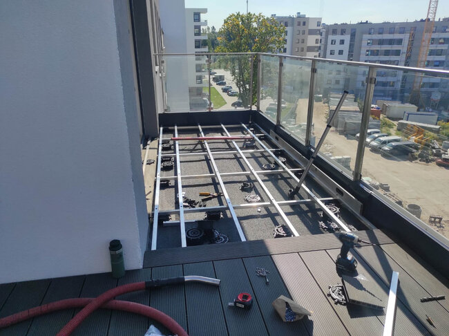 Instalación de terraza de balcón en progreso mostrando estructura de vigas de aluminio, plots soporte regulables y tablas compuestas.