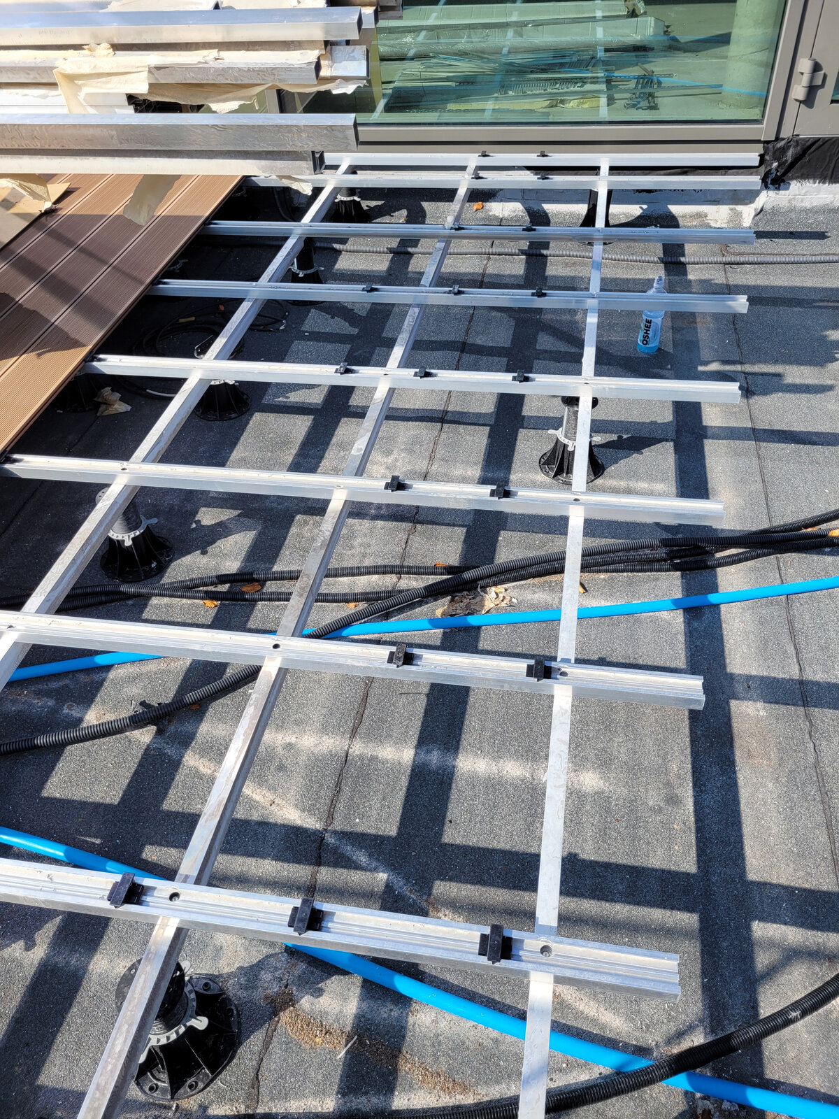 Estructura de vigas de aluminio montada sobre plots soporte regulables, tendida sobre tuberías y cableado bajo una terraza elevada.