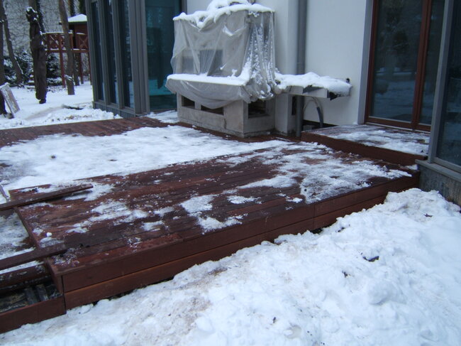 Terraza de madera oscura parcialmente cubierta de nieve derritiéndose frente a una casa en invierno.