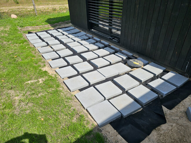 Baldosas cuadradas de hormigón colocadas sobre tela anti-malezas para preparar la base de una terraza exterior.
