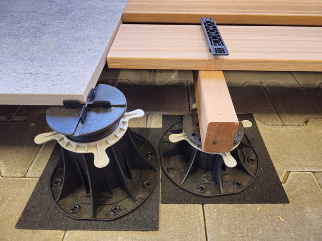 Plots Soporte Regulables que sostienen baldosas de gres porcelánico exteriores y vigas de terraza de madera.