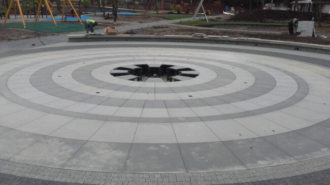 Instalación circular de suelo elevado de baldosas alrededor de una fuente seca exterior apoyada sobre soportes regulables.